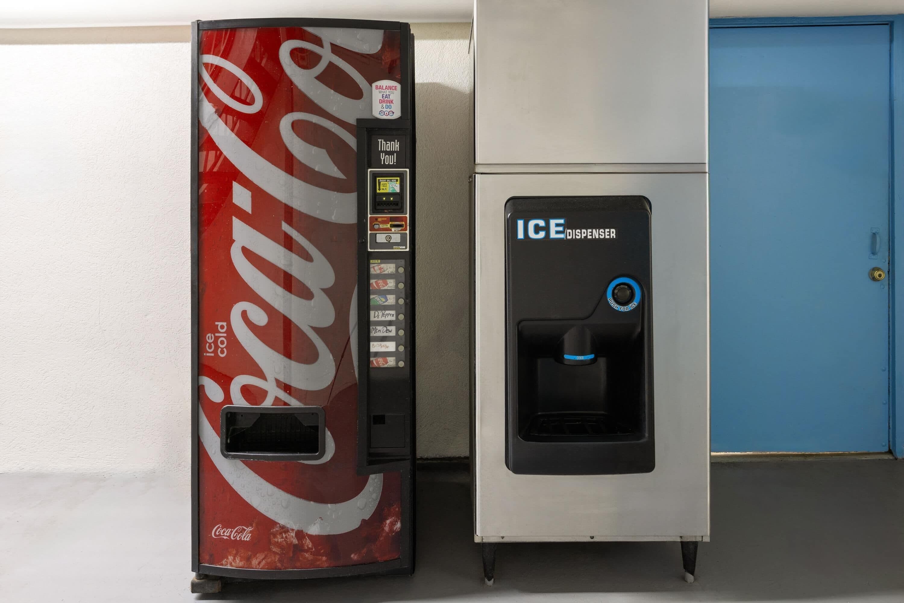 A Coca-Cola vending machine and an ICE dispenser next to a blue door.