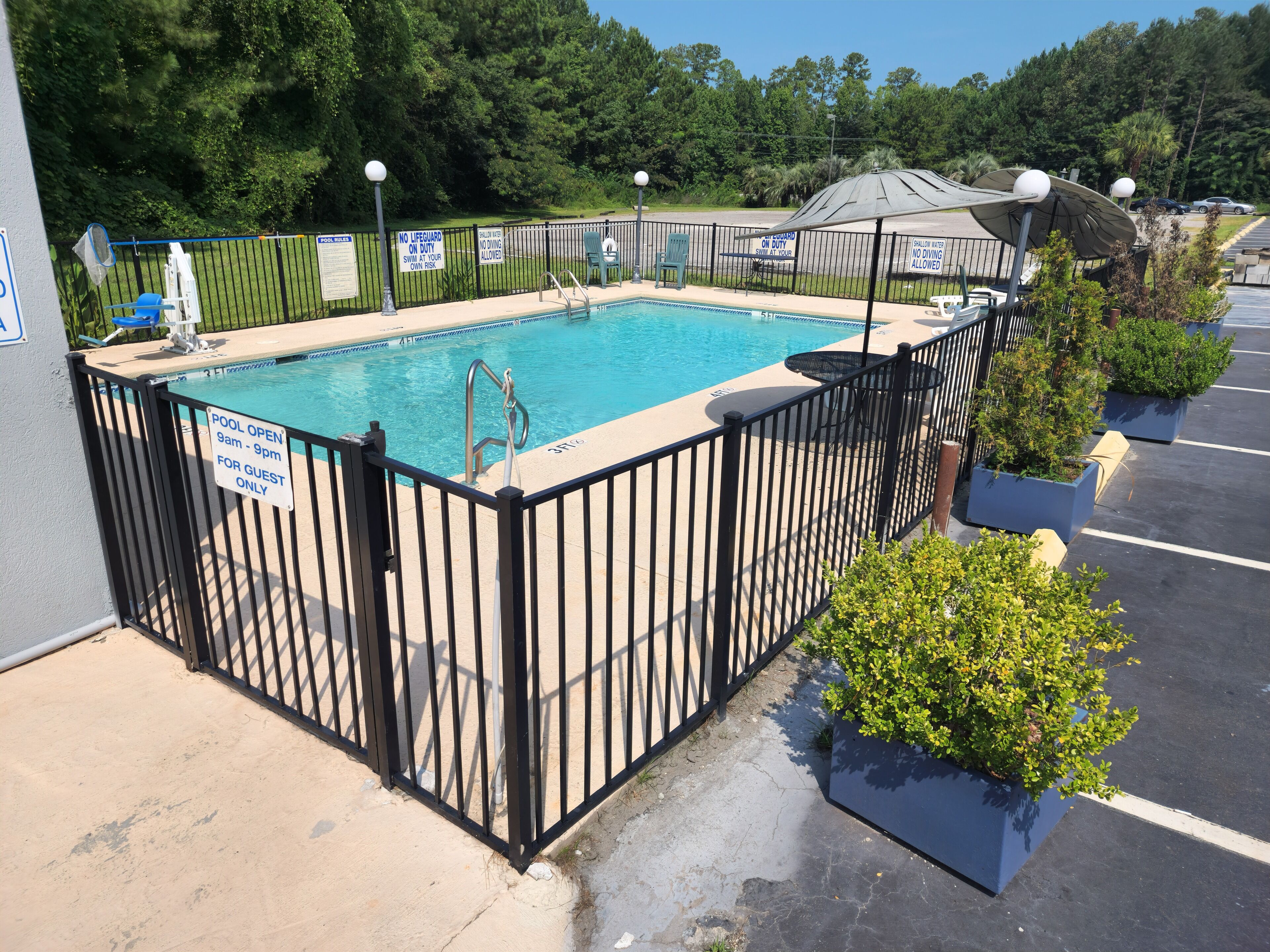 A swimming pool with a black fence, surrounded by potted plants and a parking lot.