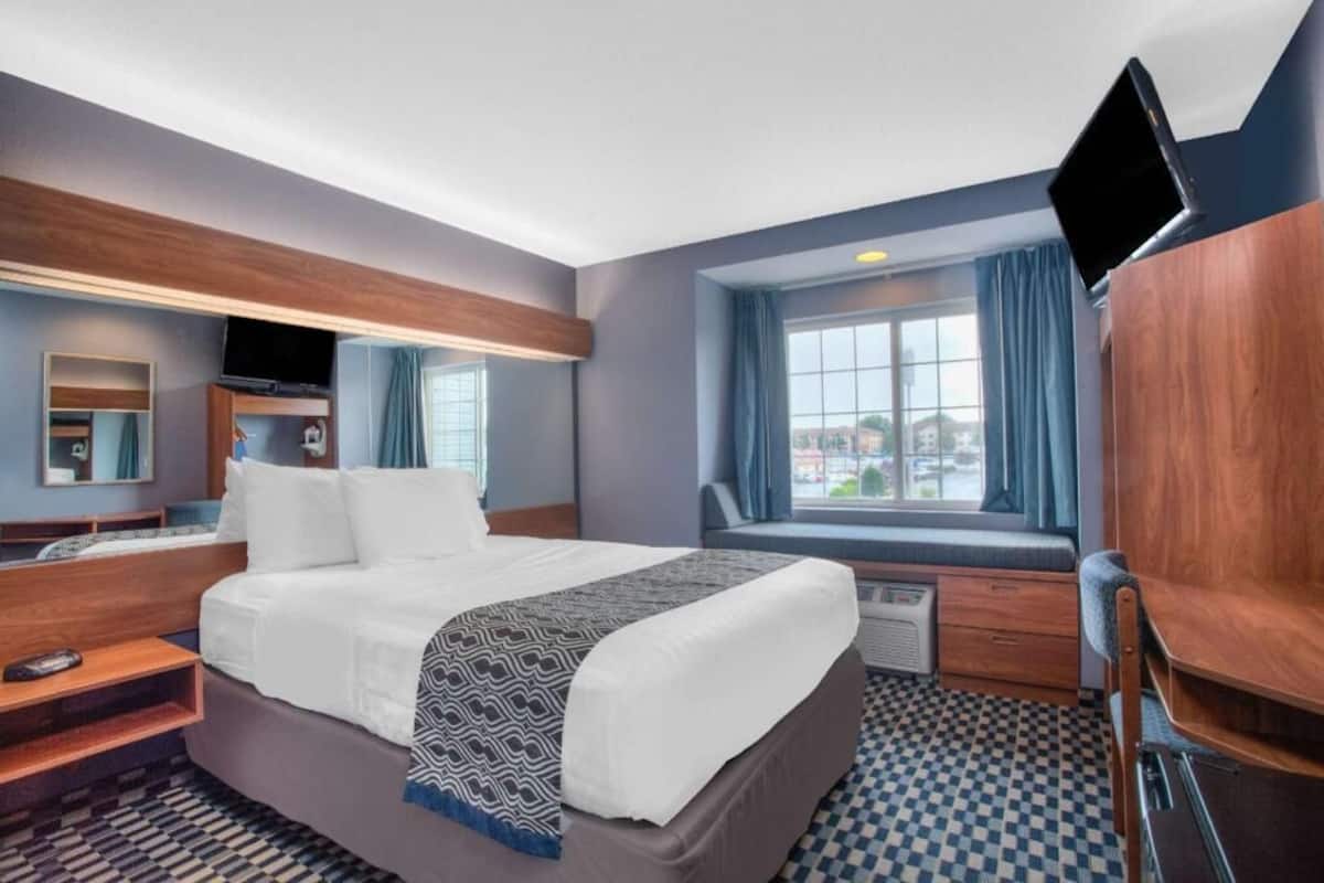 A hotel room with a bed, a TV, a window with curtains, a desk, and a chair.