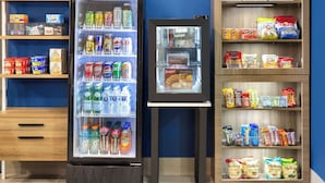 A vending machine stocked with beverages and snacks, a refrigerator with food items, and shelves with packaged goods. - Microtel Inn & Suites by Wyndham Dover (Dover)