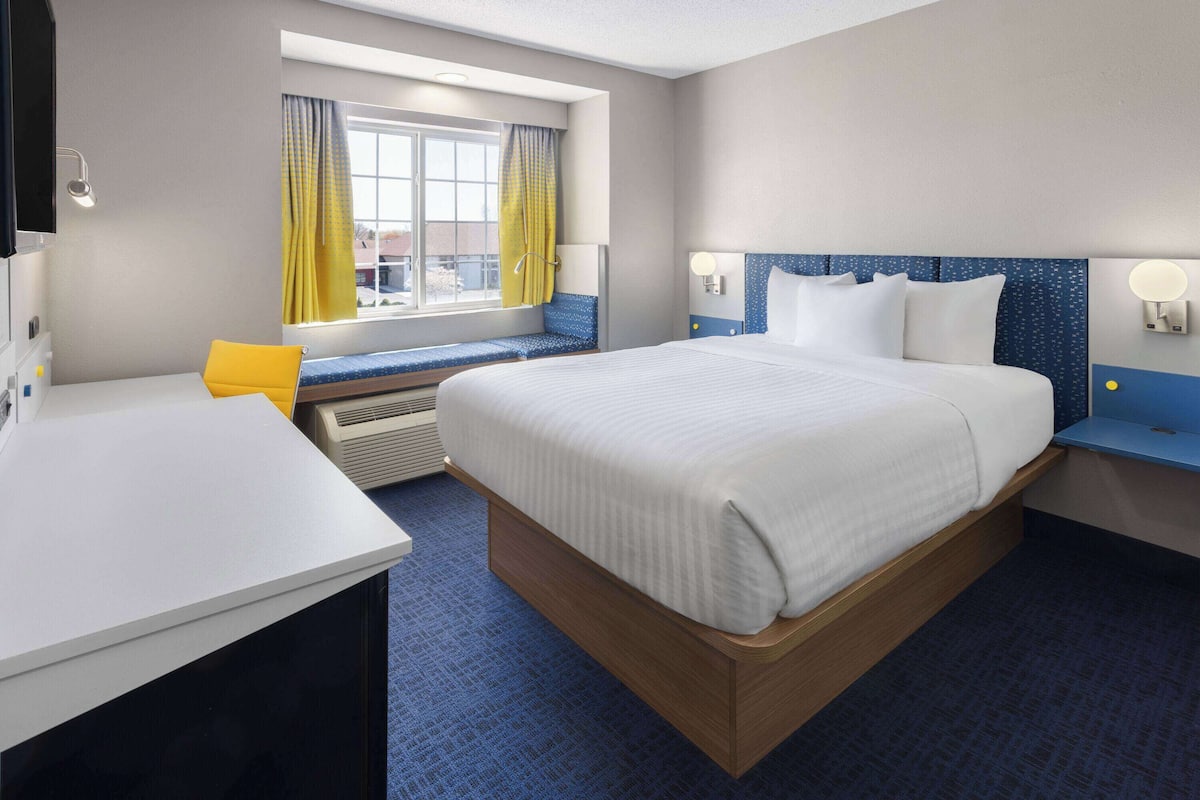 A hotel room with a large bed, a desk, a window with yellow curtains, and a view of the outside.