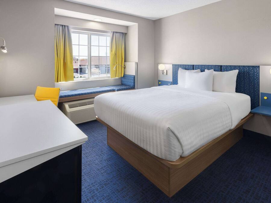 A hotel room with a large bed, a desk, a window with yellow curtains, and a view of the outside.
