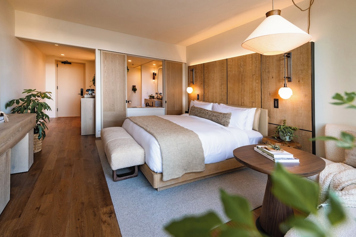 A modern hotel room with a large bed, bedside tables, a seating area, and a bathroom visible through an open door.