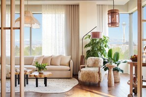 A modern living room with a sofa, armchair, and coffee table, large windows with a view of a tower, and various plants.