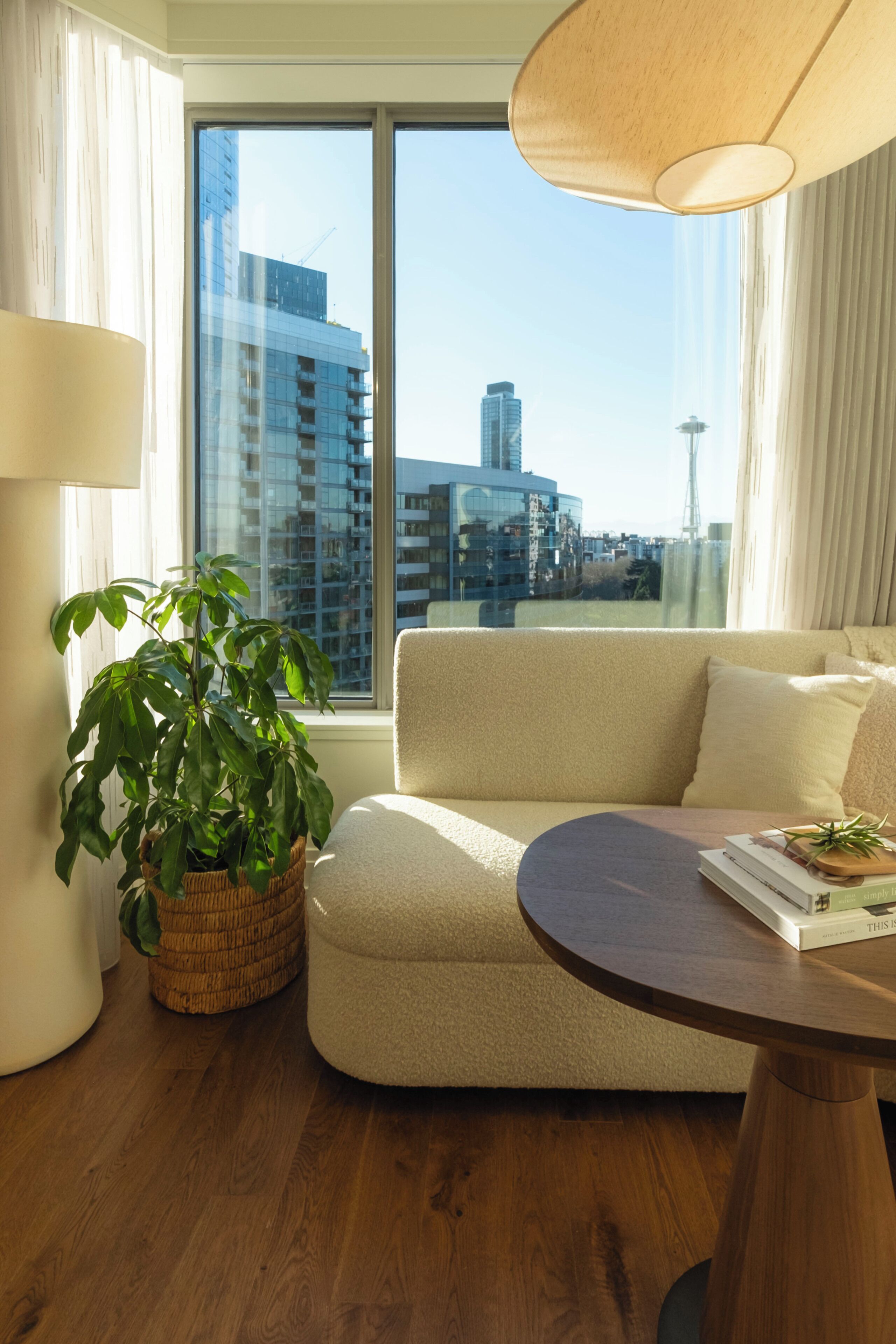 Una sala de estar moderna con un sofá, una mesa redonda de madera, una planta en maceta y vistas de un paisaje urbano a través de amplios ventanales.