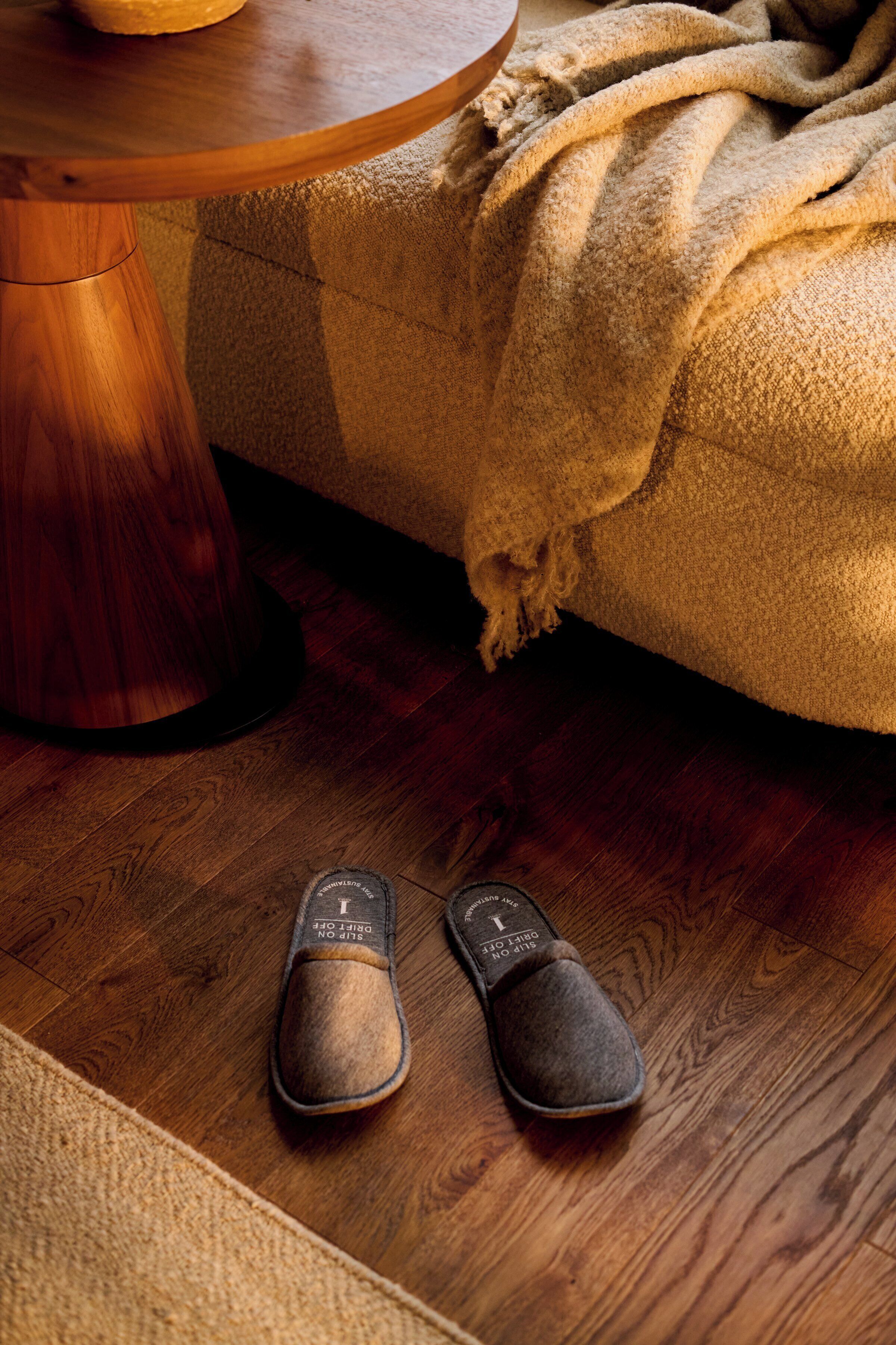 Un par de pantuflas sobre un piso de madera, junto a una mesa redonda de madera y un sofá beige con una manta.
