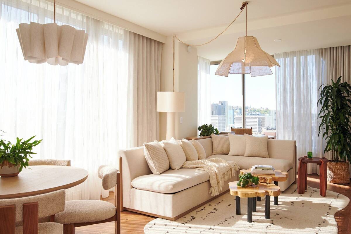 A modern living room with a beige sectional sofa, a round wooden dining table, and a large window with sheer curtains.