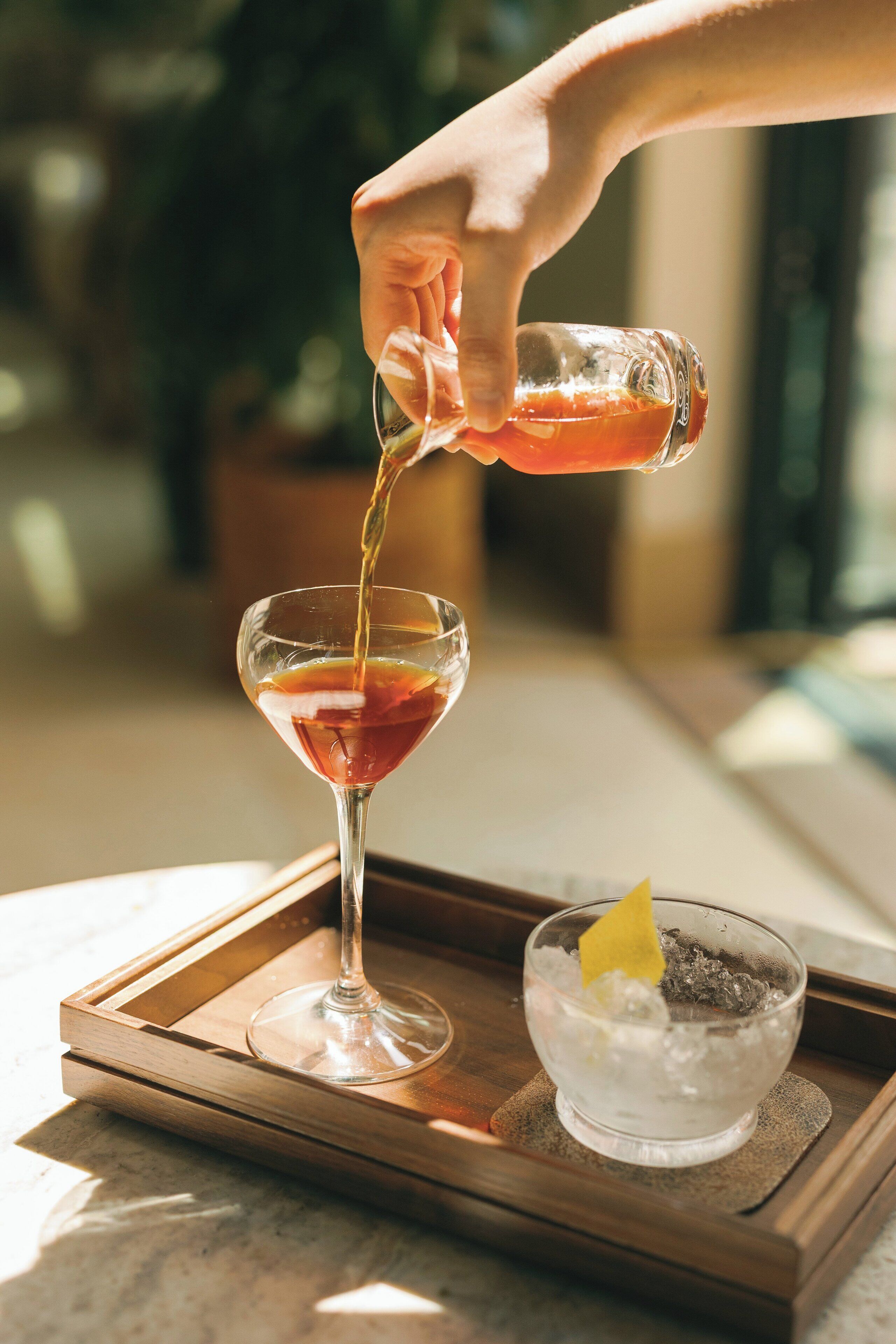 A hand pouring a drink into a martini glass on a tray.