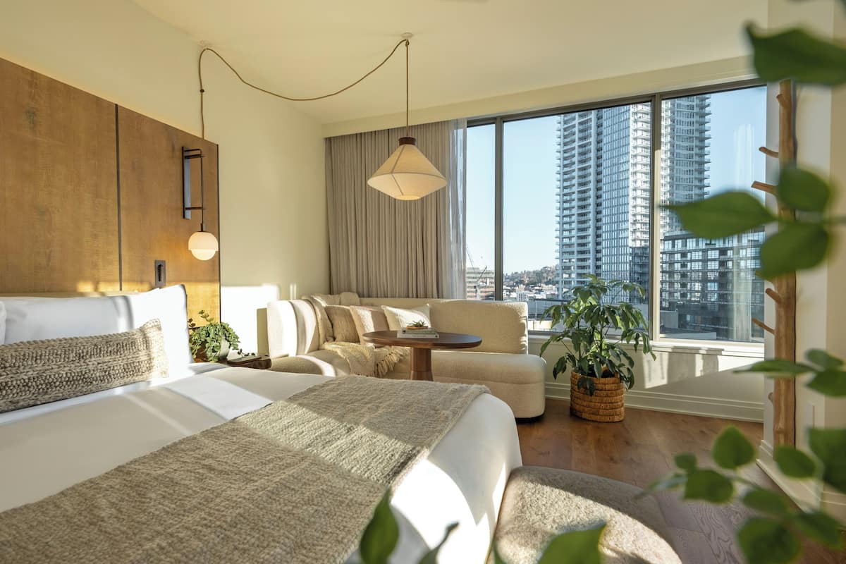 A modern hotel room with a large bed, a sofa, a small table, and a view of tall buildings.