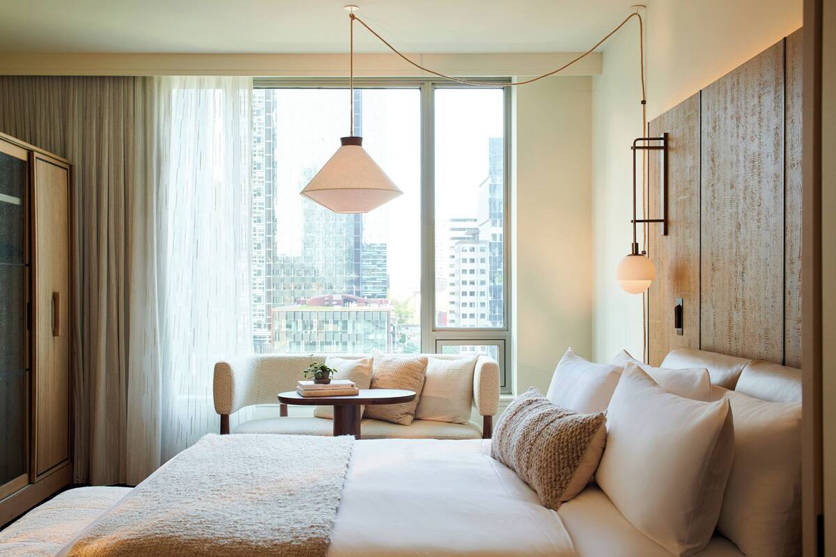A hotel room with a large bed, a sofa, a small table, and a city view through large windows.