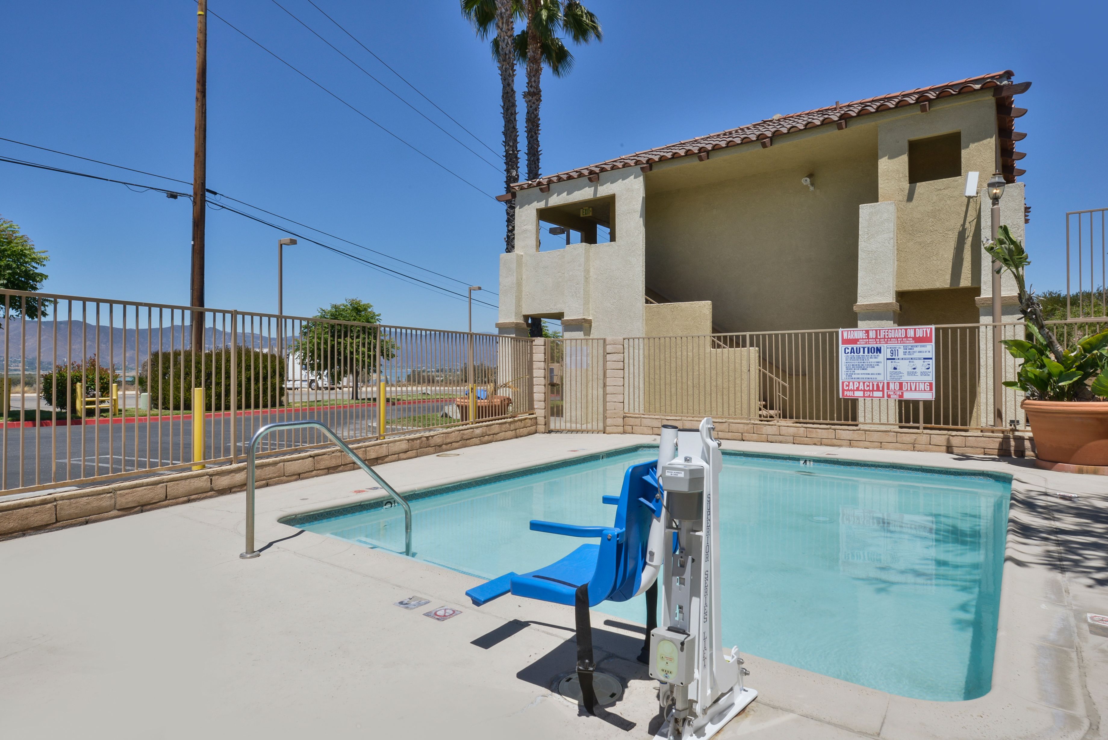 A swimming pool with a lifeguard chair and a lifeguard stand, surrounded by a fence and a building with a sign indicating no diving.