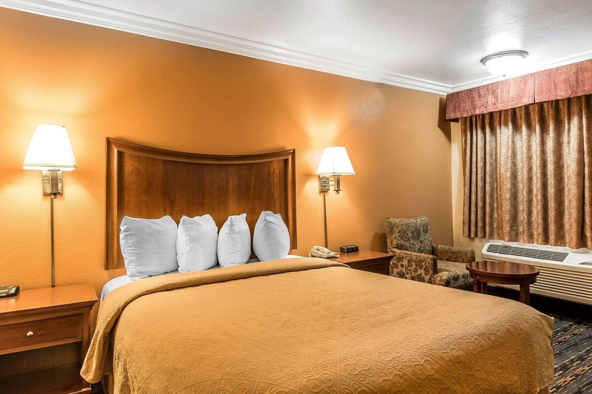 A hotel room with a large bed, two bedside lamps, a chair, a desk, and an air conditioning unit.