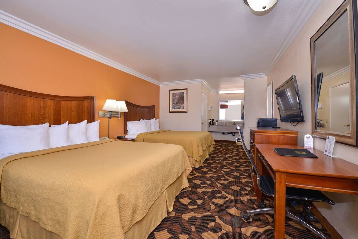 A hotel room with two beds, a desk with a computer, a television, and a bathroom visible in the background.