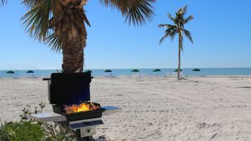 Een strand met een barbecue, palmbomen en strandstoelen onder parasols.