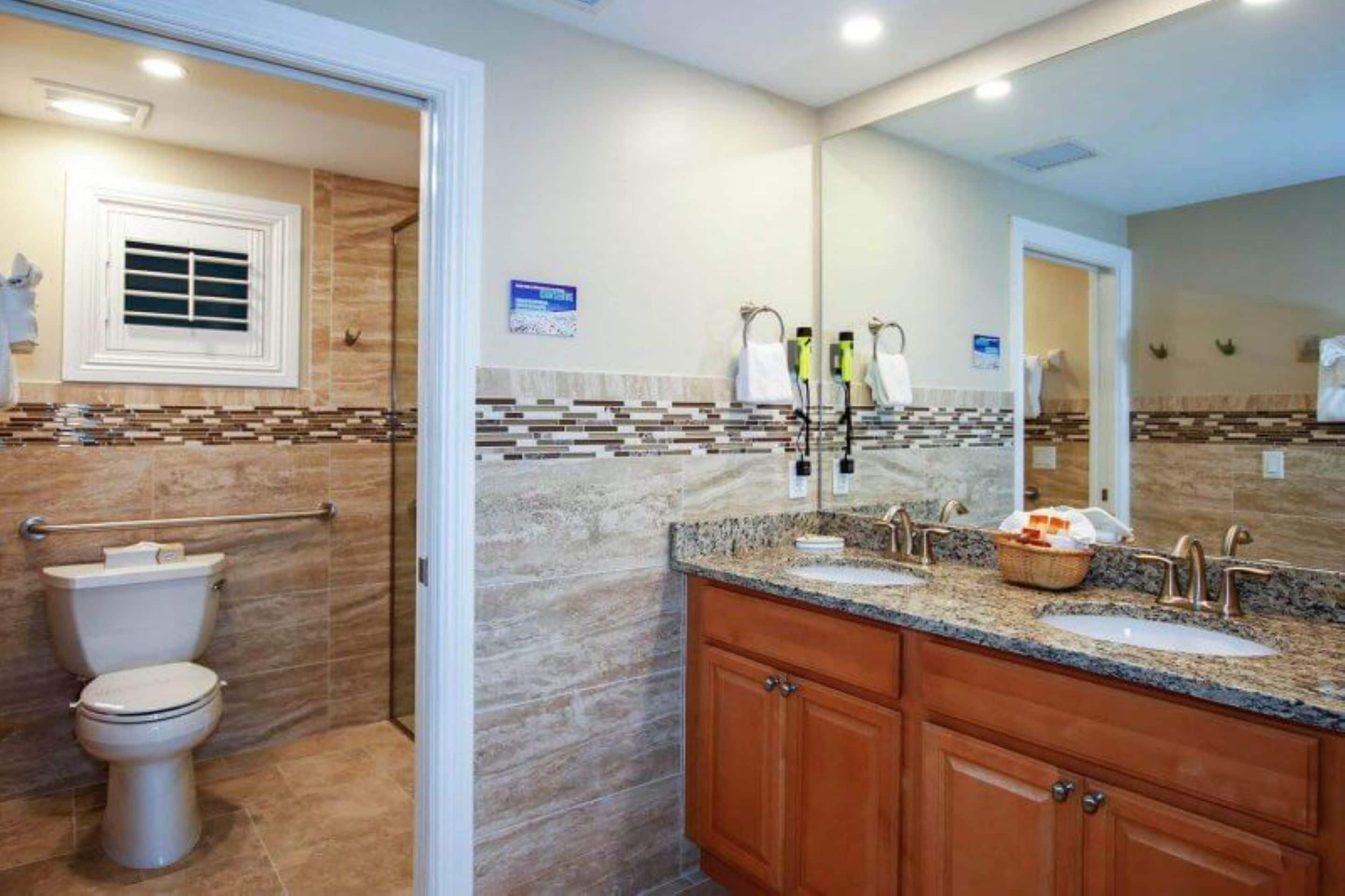 Un baño moderno con ducha, inodoro y tocador con doble lavamanos y un espejo grande.