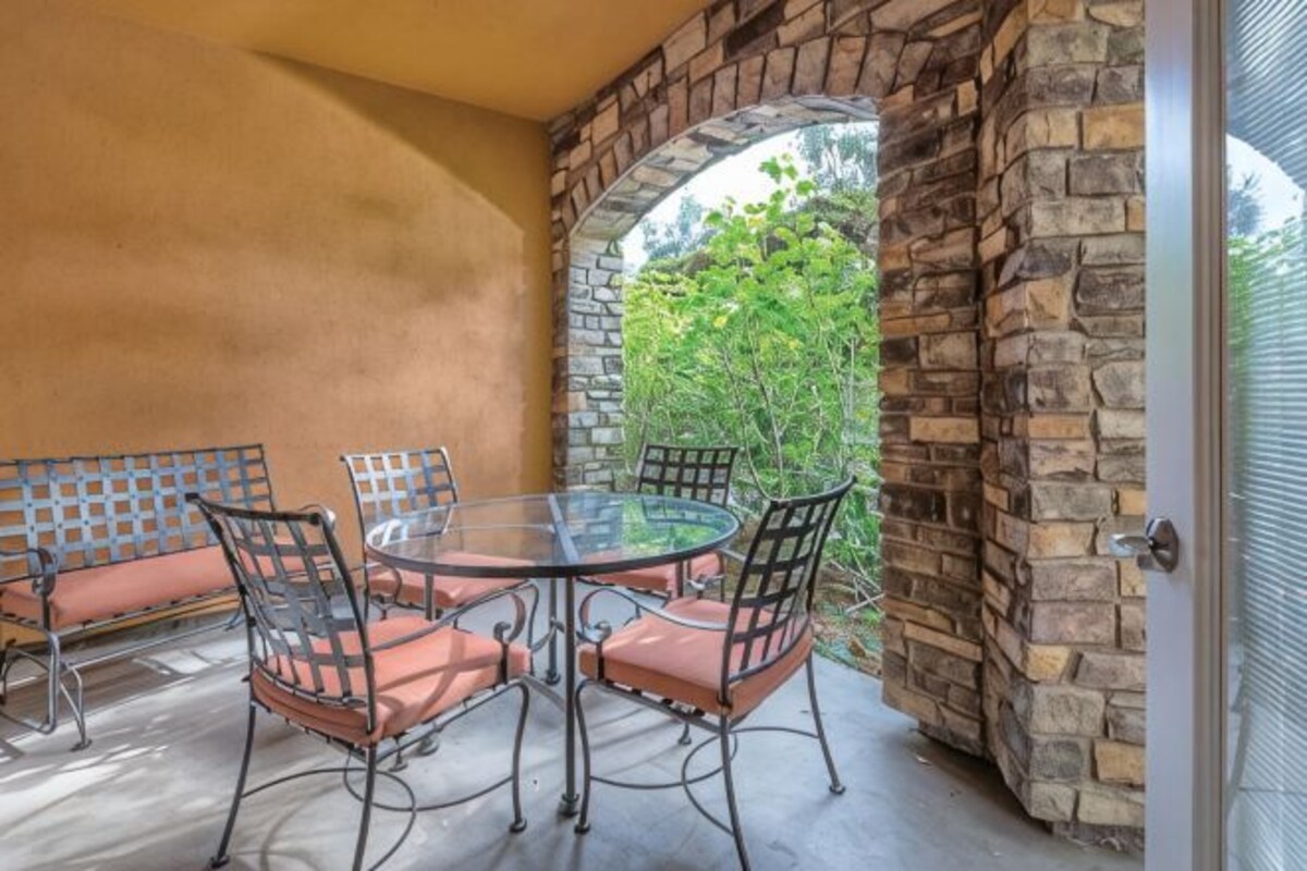 A patio with a round table and chairs, a stone wall, and a view of greenery.
