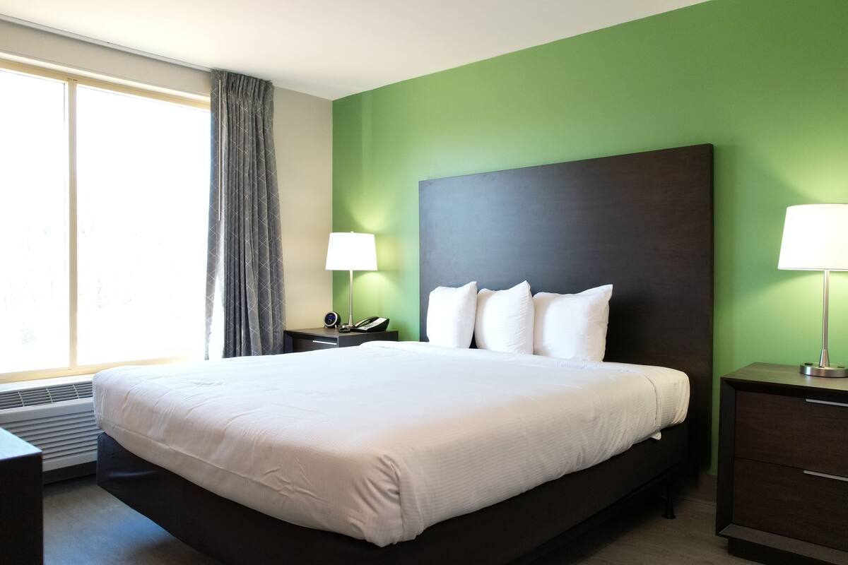Une chambre d’hôtel avec un grand lit, un bureau, une chaise, deux tables de chevet avec des lampes, une fenêtre avec des rideaux et un mur vert.