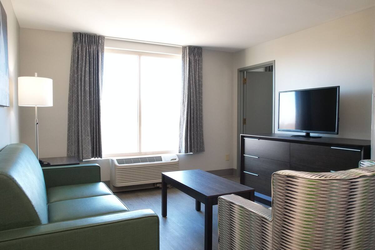Une chambre d’hôtel de taille modeste, avec un canapé vert, une petite table, un téléviseur posé sur un meuble et une fenêtre avec des rideaux.