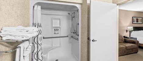 A hotel bathroom with a walk-in shower, a towel rack with folded towels, a hand dryer, and a small shelf with toiletries.