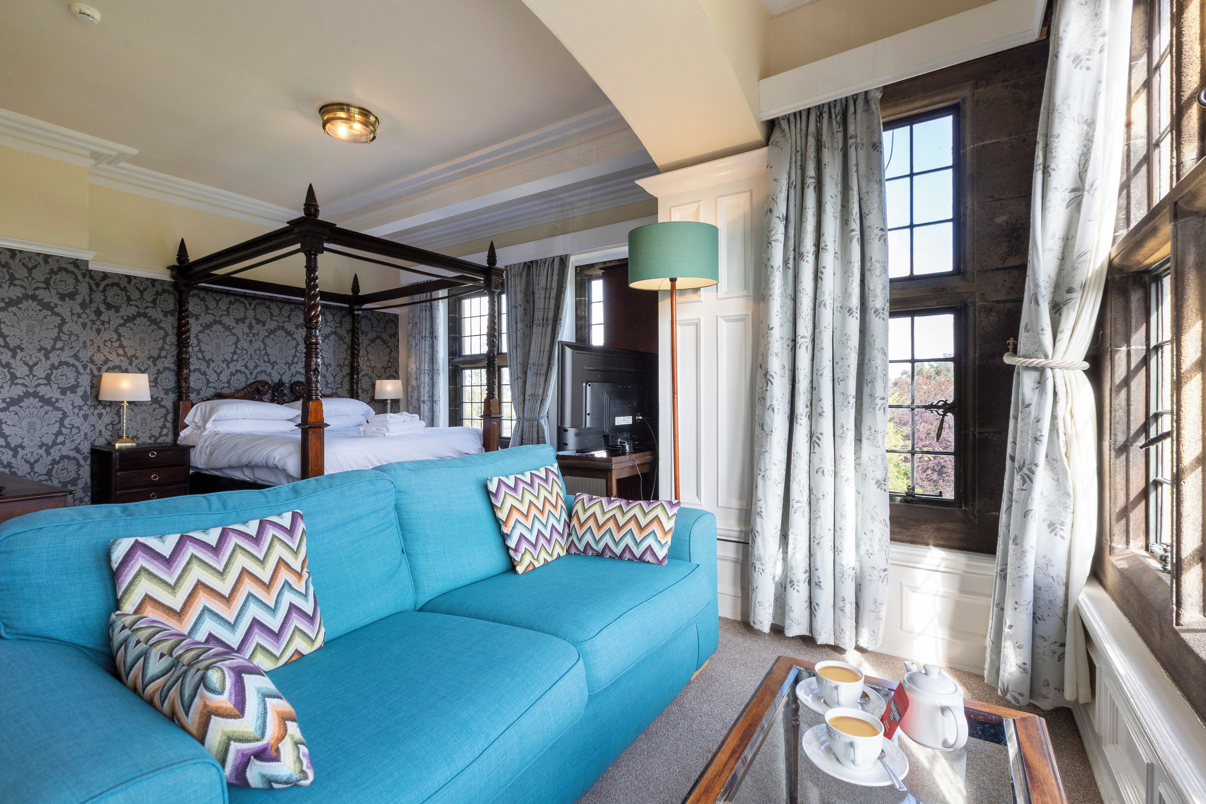 A hotel room with a four-poster bed, a blue sofa with patterned cushions, a coffee table with cups and a teapot, and large windows with curtains.