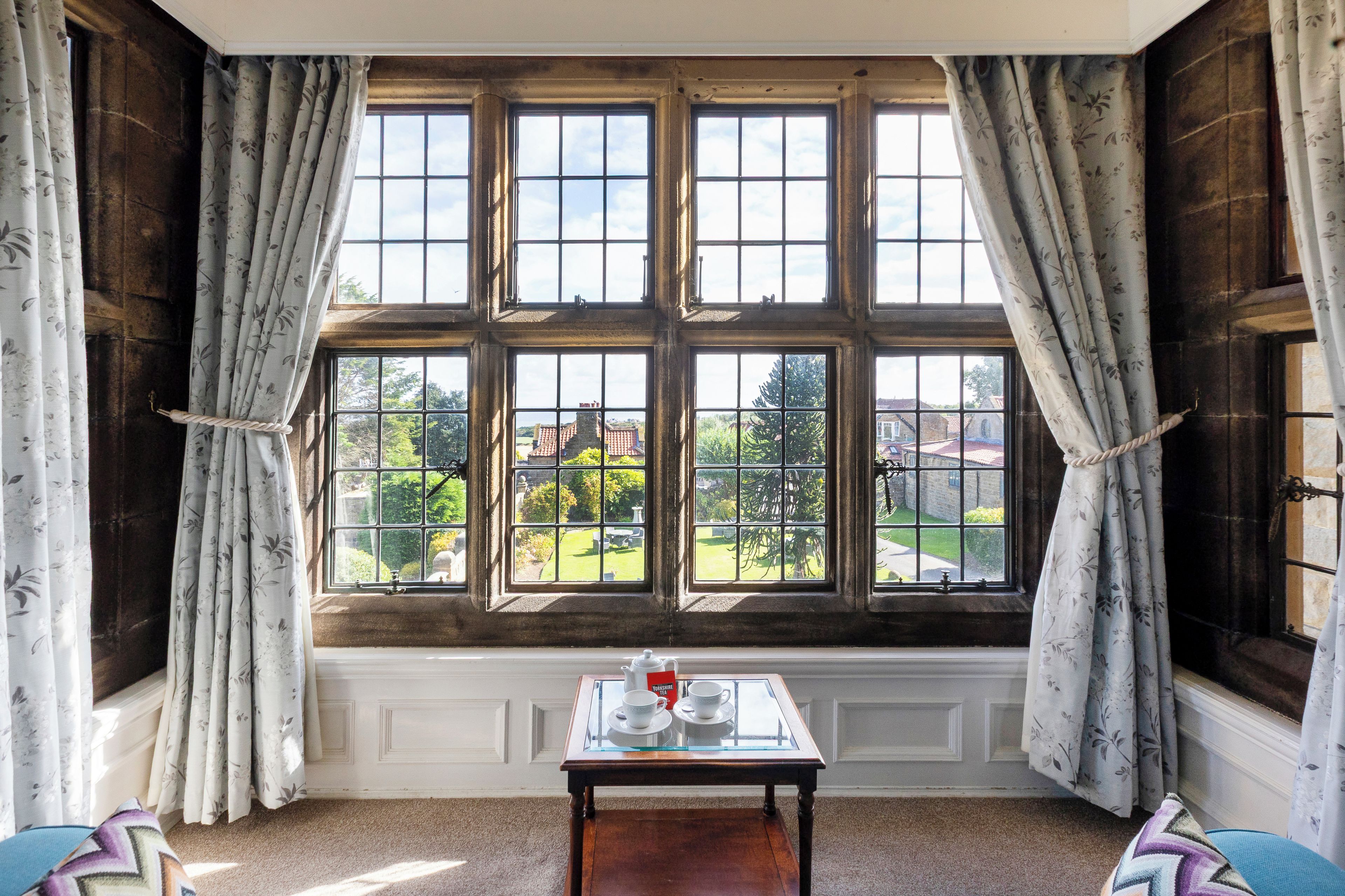 A room with a large window, a small table with a teapot and cups, and floral curtains.