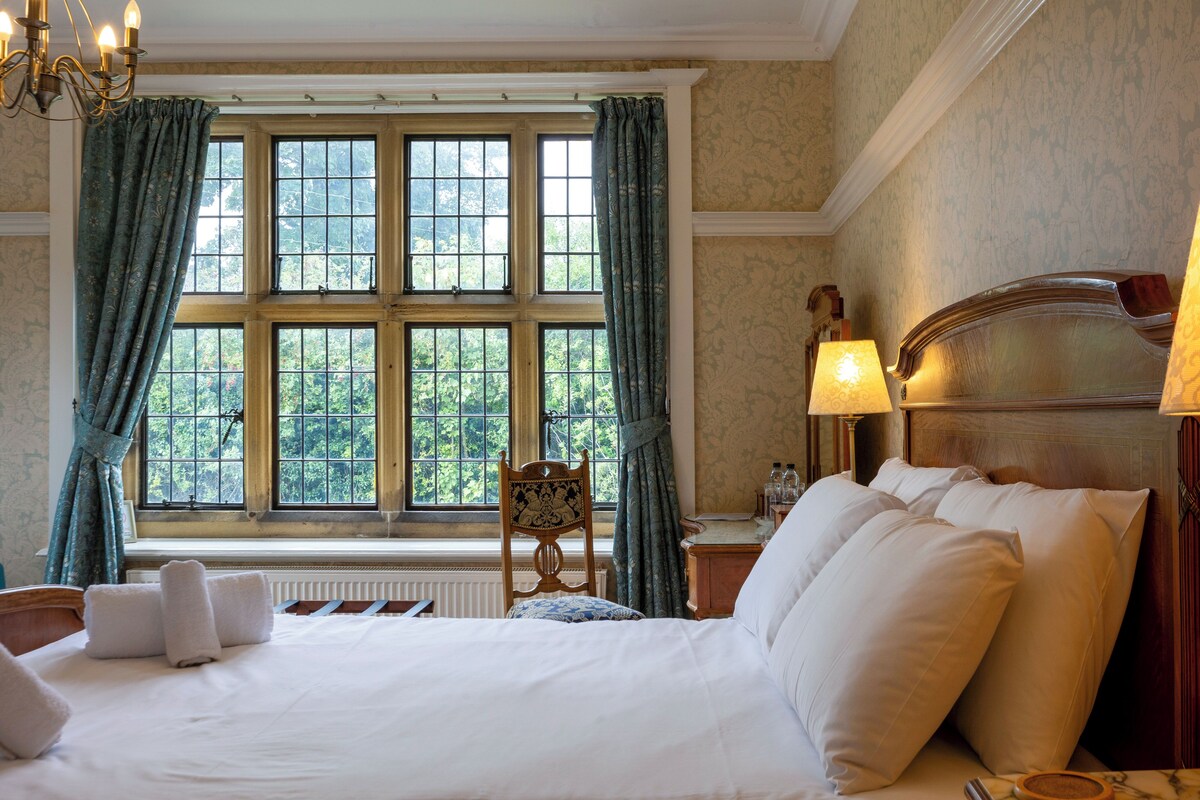 Ett hotellrum med ett stort fönster, en säng med vita sängkläder, en trästol och två sänglampor.