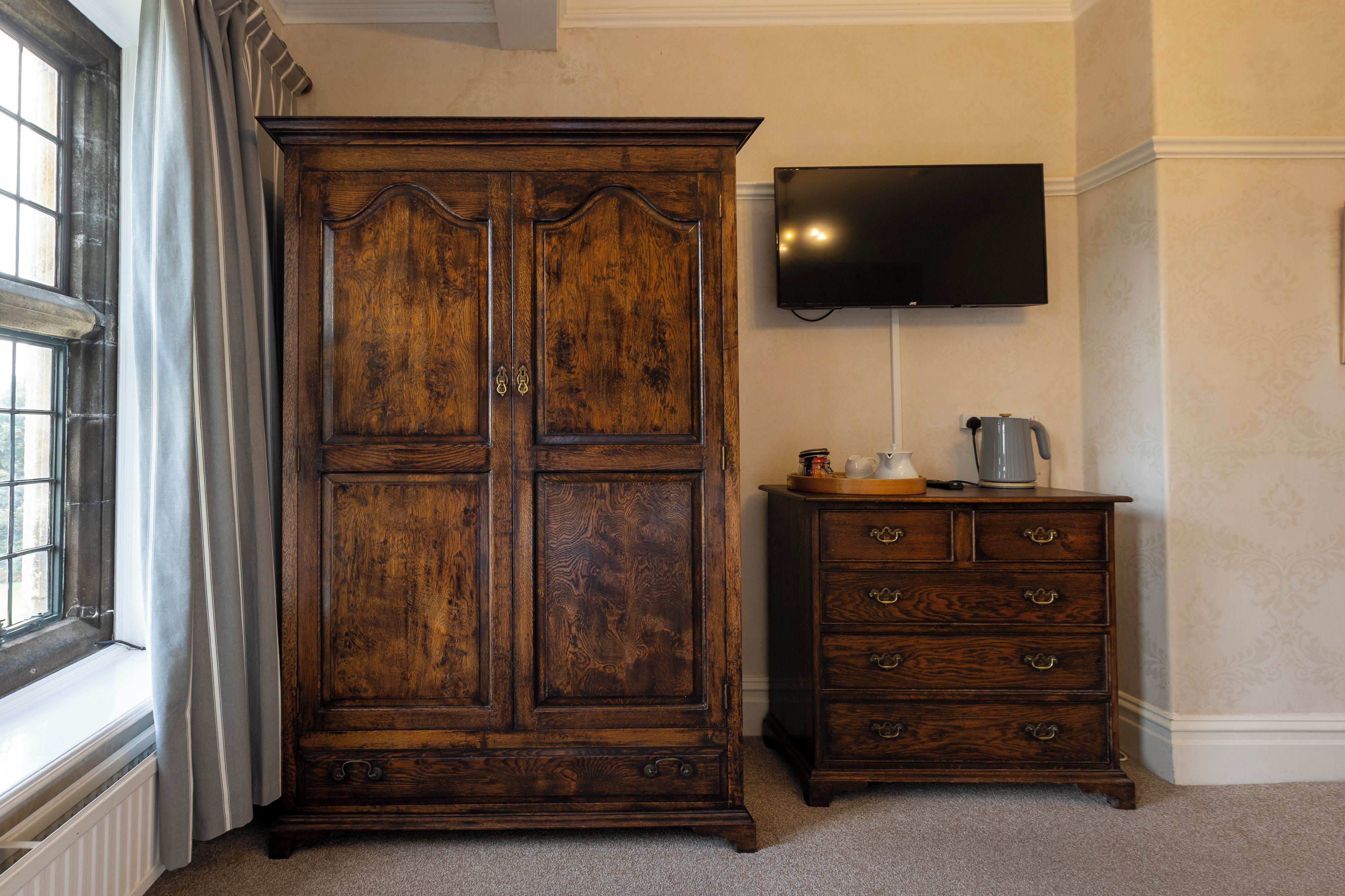 Una habitación con un armario de madera grande, un pequeño tocador con cajones, un televisor de pantalla plana en la pared y una ventana con cortinas.