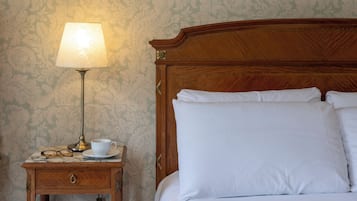 Una cama bien hecha con sábanas blancas, cabecera de madera y mesita de noche con lámpara, taza y vasos.