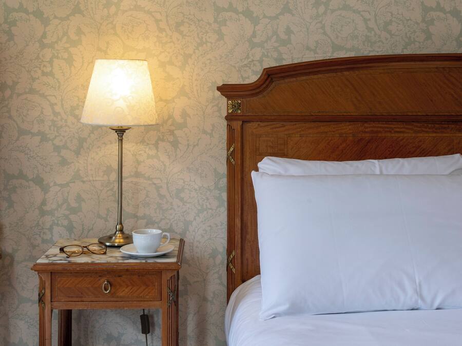 A neatly made bed with white linens, a wooden headboard, and a bedside table with a lamp, cup, and glasses.