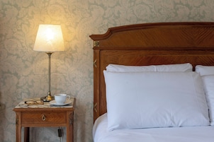 Una cama bien hecha con sábanas blancas, cabecera de madera y mesita de noche con lámpara, taza y vasos.