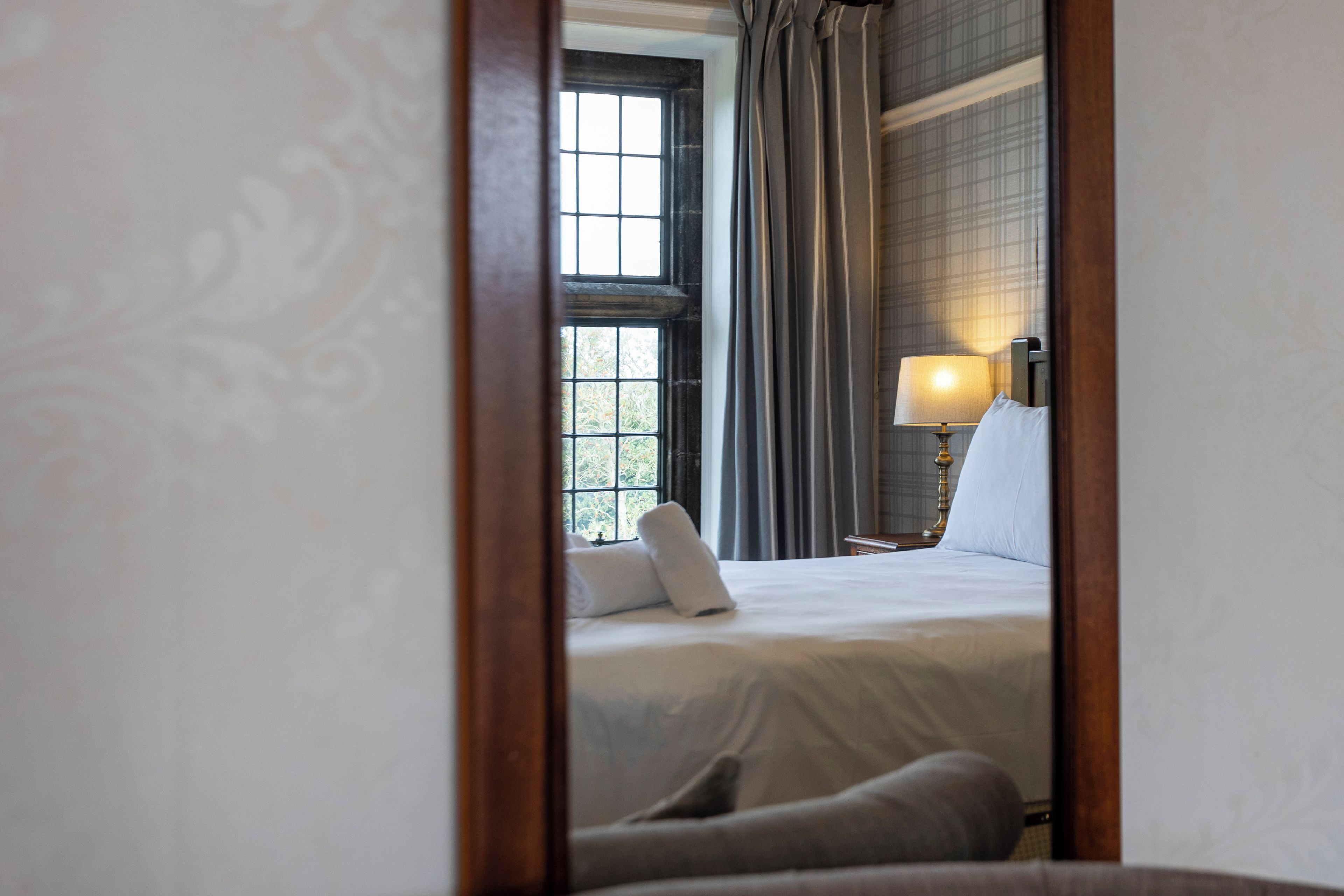 Quarto de hotel com cama, mesa de cabeceira com abajur e janela com cortinas.