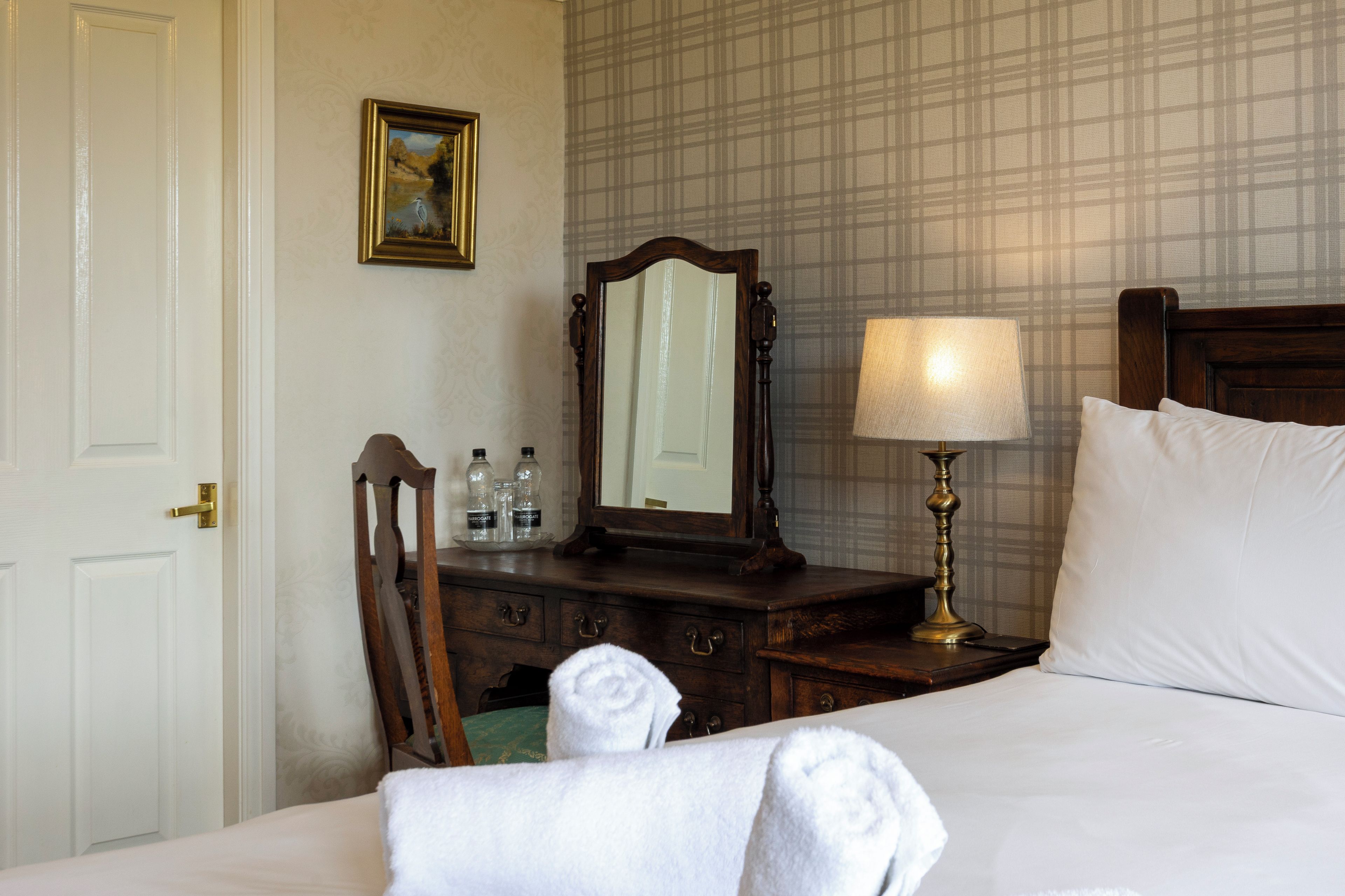 Een hotelkamer met een bed, nachtkastje, spiegel, lamp, dressoir en een deur.