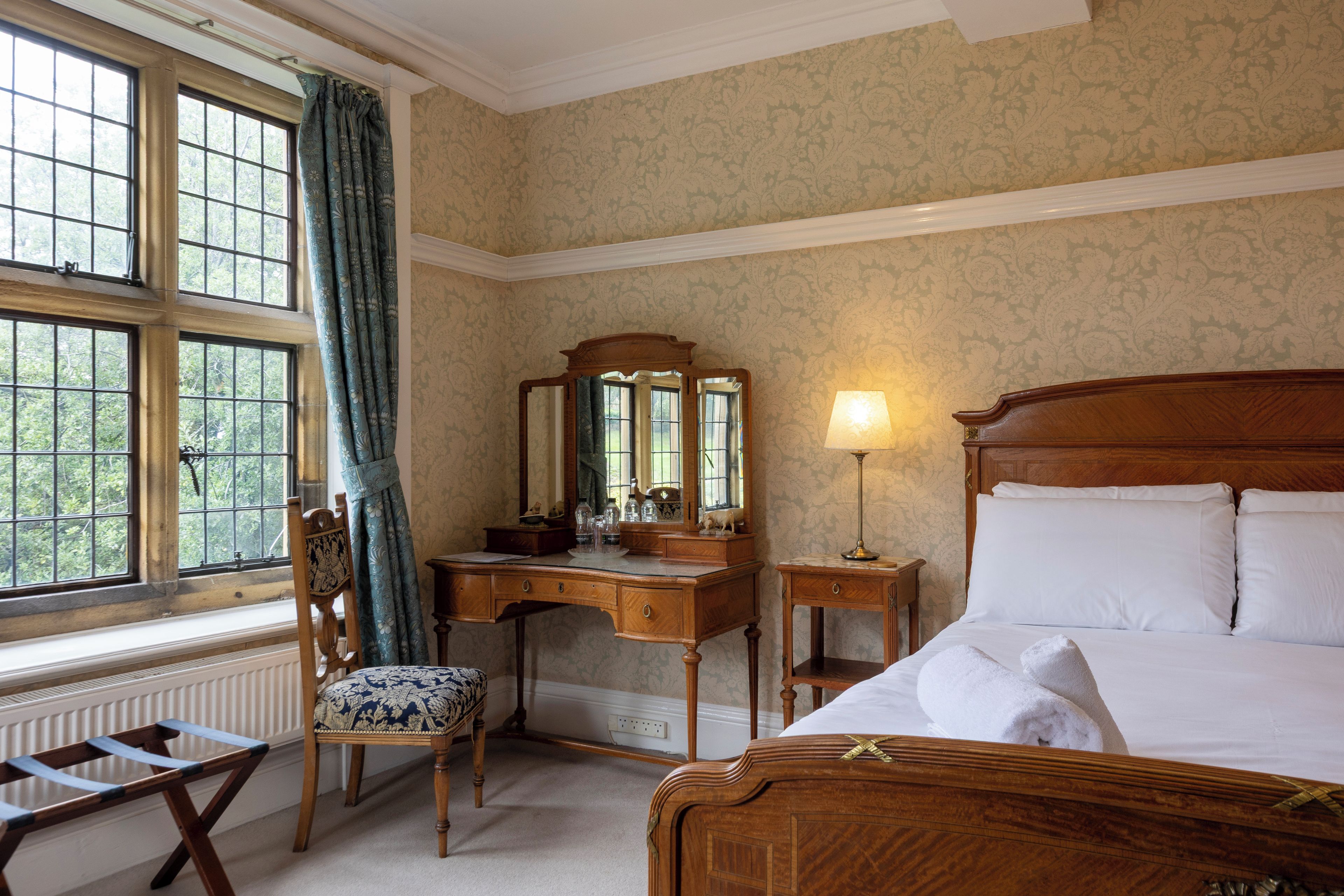A hotel room with a wooden bed, a desk with a mirror, a chair, and a window with curtains.