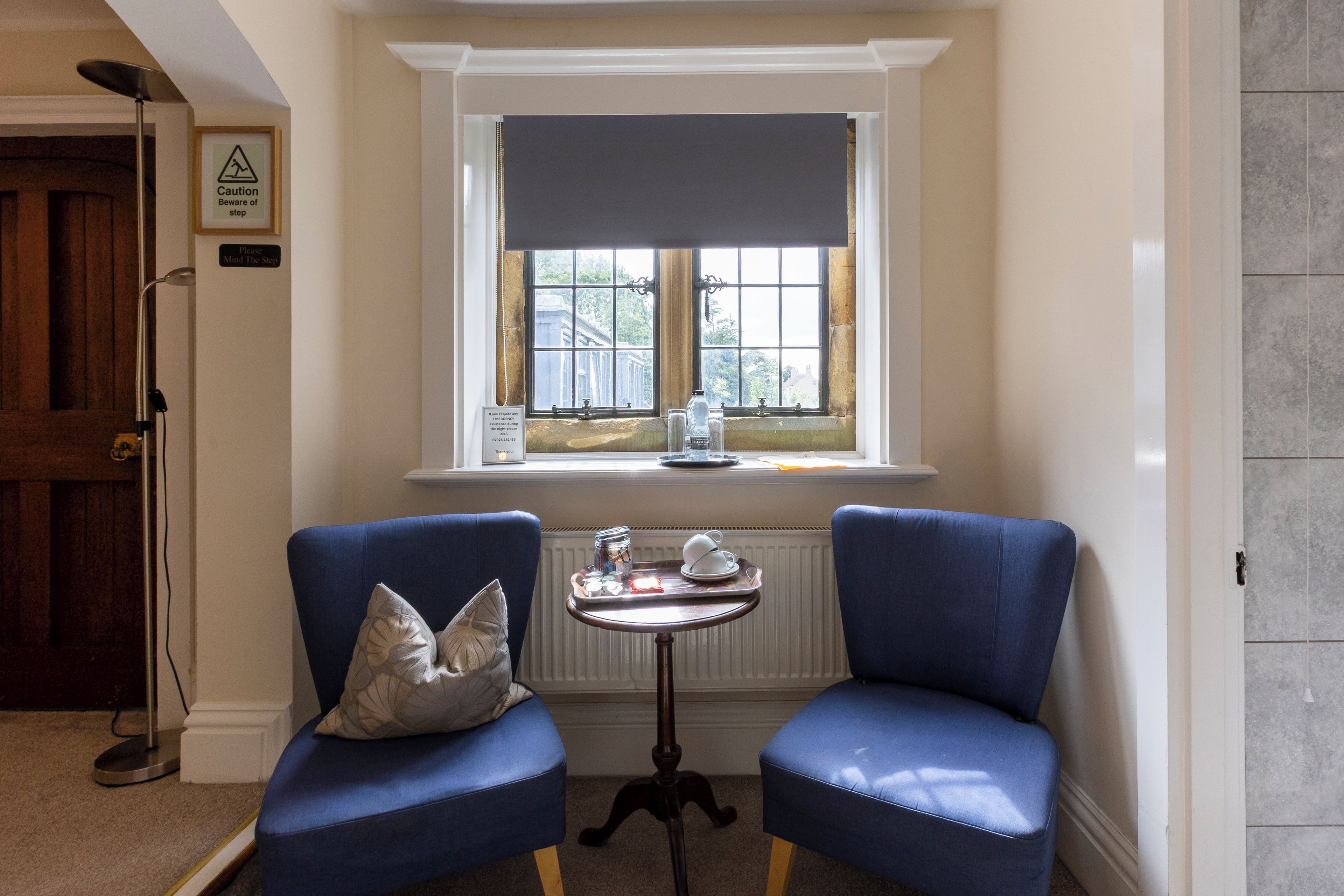 Une pièce d’angle avec deux fauteuils bleus, une petite table sur laquelle se trouvent une théière et des tasses, et une fenêtre donnant sur des arbres.