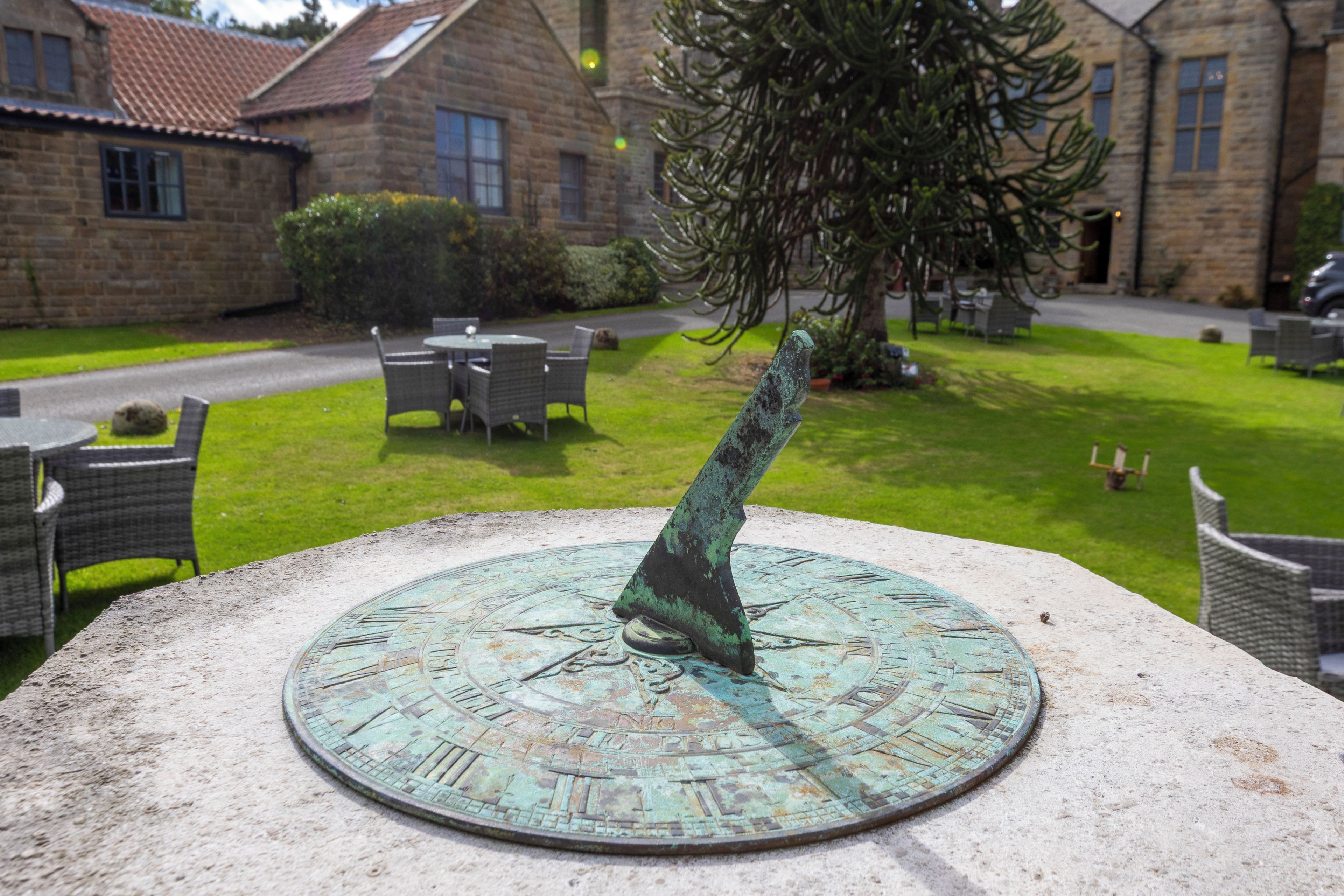 A sundial with Roman numerals and a gnomon in a courtyard with stone buildings and outdoor furniture.