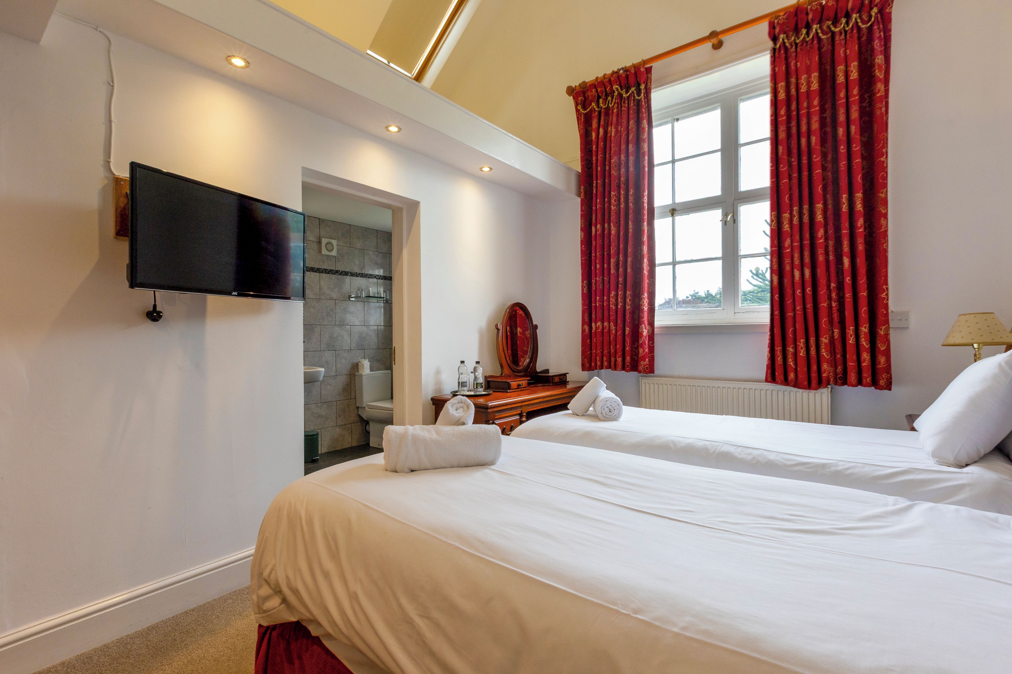 A hotel room with two beds, a television, a bathroom, and red curtains.
