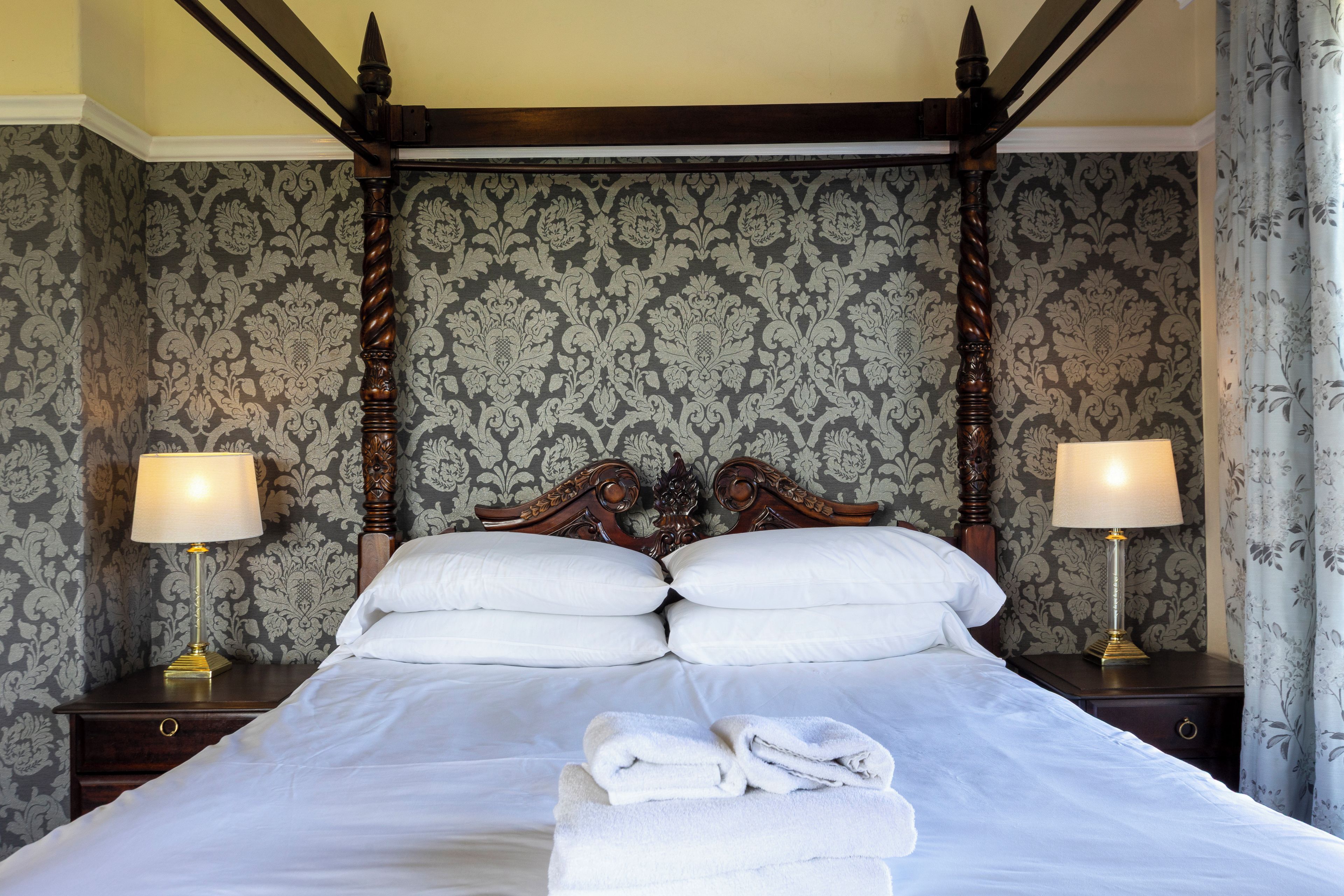 A four-poster bed with white linens and pillows, flanked by two bedside tables with lamps, set against a patterned wallpaper backdrop.