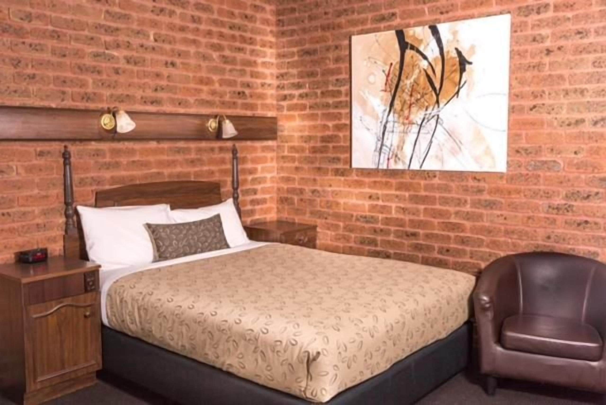 A bedroom with a brick wall, a bed with a patterned bedspread, a nightstand, a lamp, and a brown leather chair.