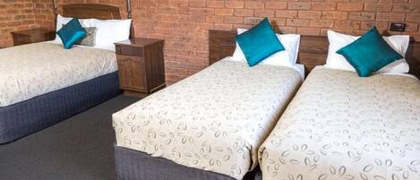 Quarto de hotel com duas camas, cada uma com colcha estampada e almofadas verde-água. O quarto possui parede de tijolos e cabeceira de madeira.