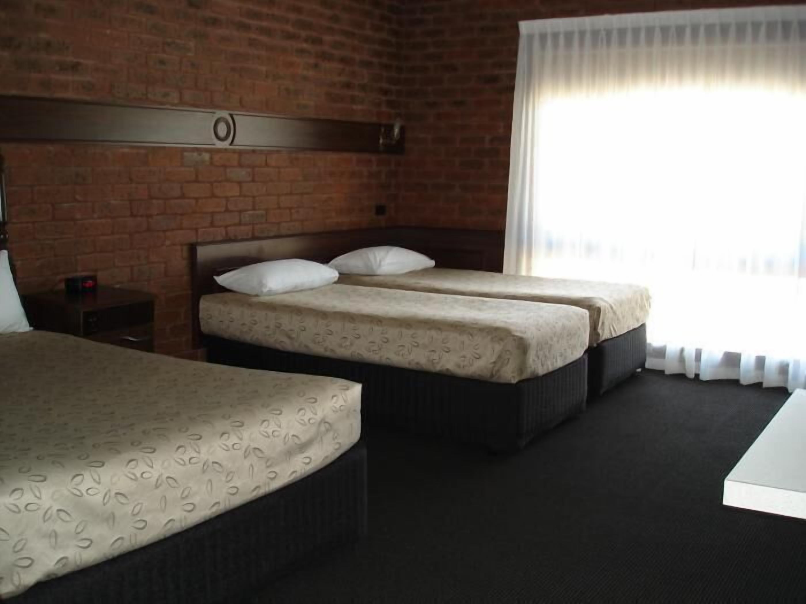 A hotel room with two single beds, a brick wall, a window with curtains, and a small bedside table with a lamp.