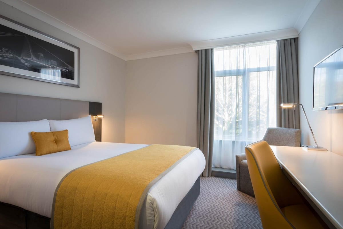 A hotel room with a large bed, a desk, a chair, a lamp, and a window with curtains.