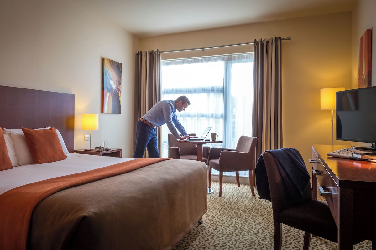 A hotel room with a large bed, a desk with a laptop, a TV, and a window with curtains.