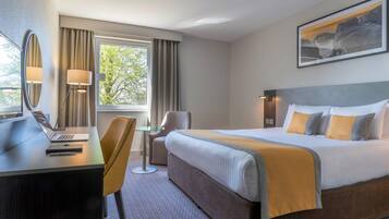 A hotel room with a bed, a desk with a laptop, a chair, a small table, and a window with curtains.