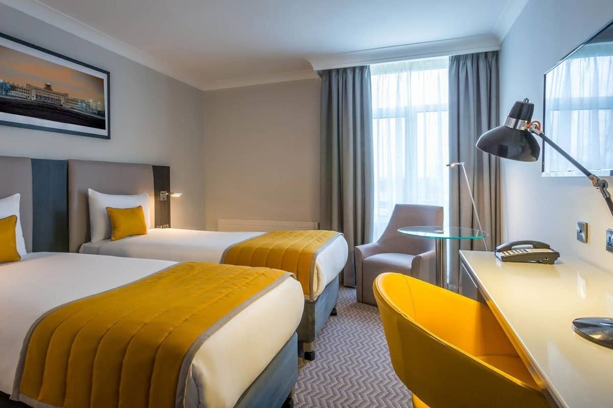 A hotel room with two beds, a desk, a chair, a lamp, and a painting on the wall.