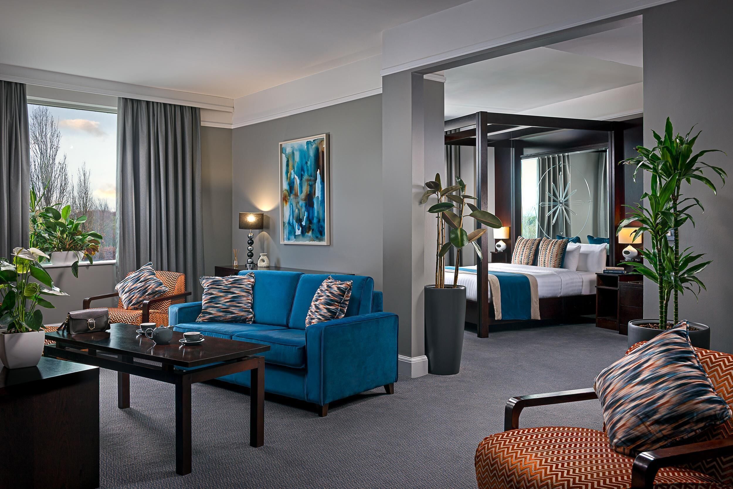 A modern hotel room with a blue sofa, a dining table, a large window with curtains, a painting on the wall, and a separate bedroom area.