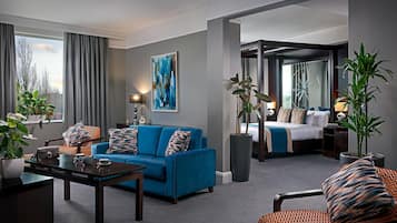 A modern hotel room with a blue sofa, a dining table, a large window with curtains, a painting on the wall, and a separate bedroom area.