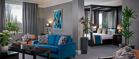A modern hotel room with a blue sofa, a dining table, a large window with curtains, a painting on the wall, and a separate bedroom area.