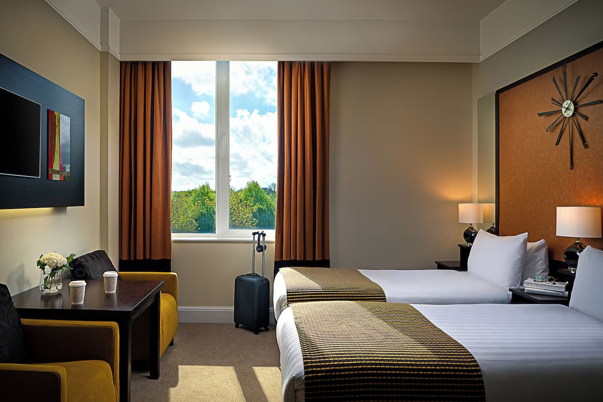 Een hotelkamer met twee bedden, een bureau, een stoel, een raam met gordijnen, een klok aan de muur en een aan de muur bevestigde televisie.