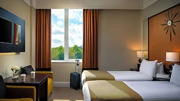 Quarto de hotel com duas camas, uma escrivaninha, uma cadeira, janela com cortinas, relógio na parede e televisão fixada na parede.
