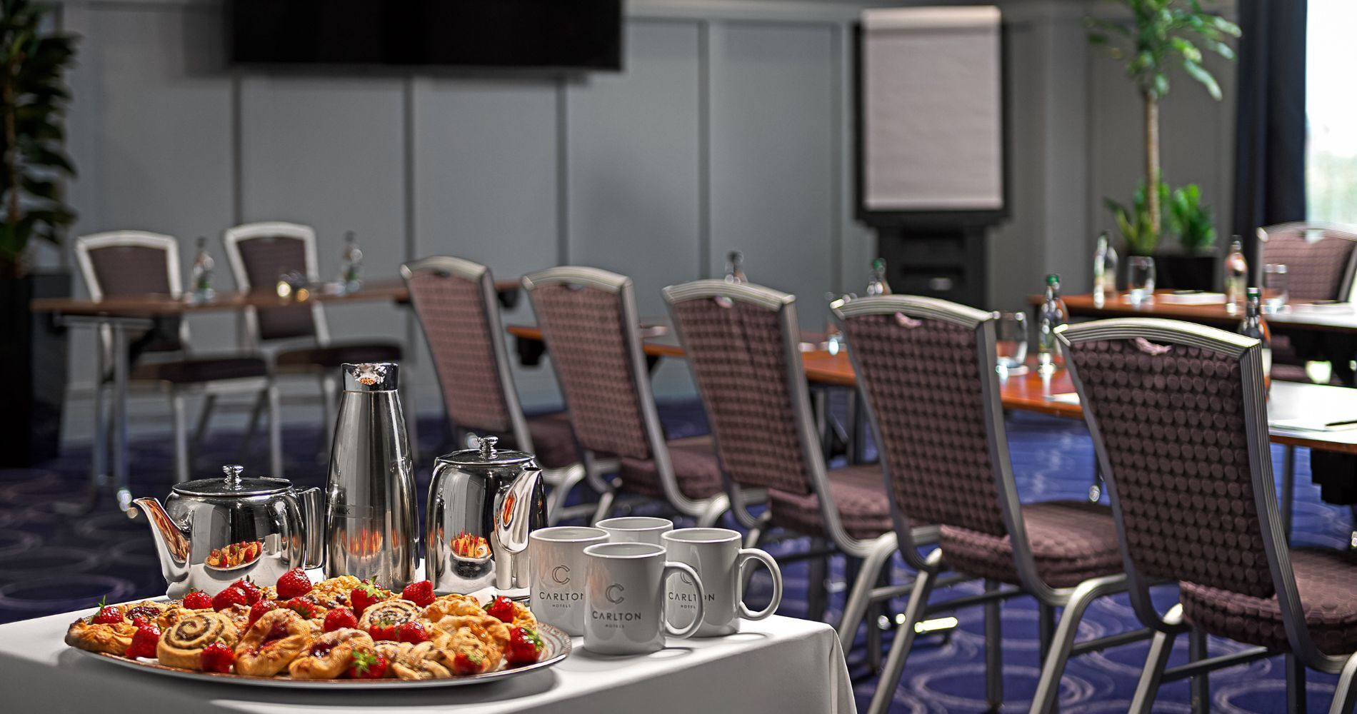 Un salón de conferencias con una mesa dispuesta para el desayuno, que incluye pasteles y tazas de café, y una pantalla de televisión al fondo.